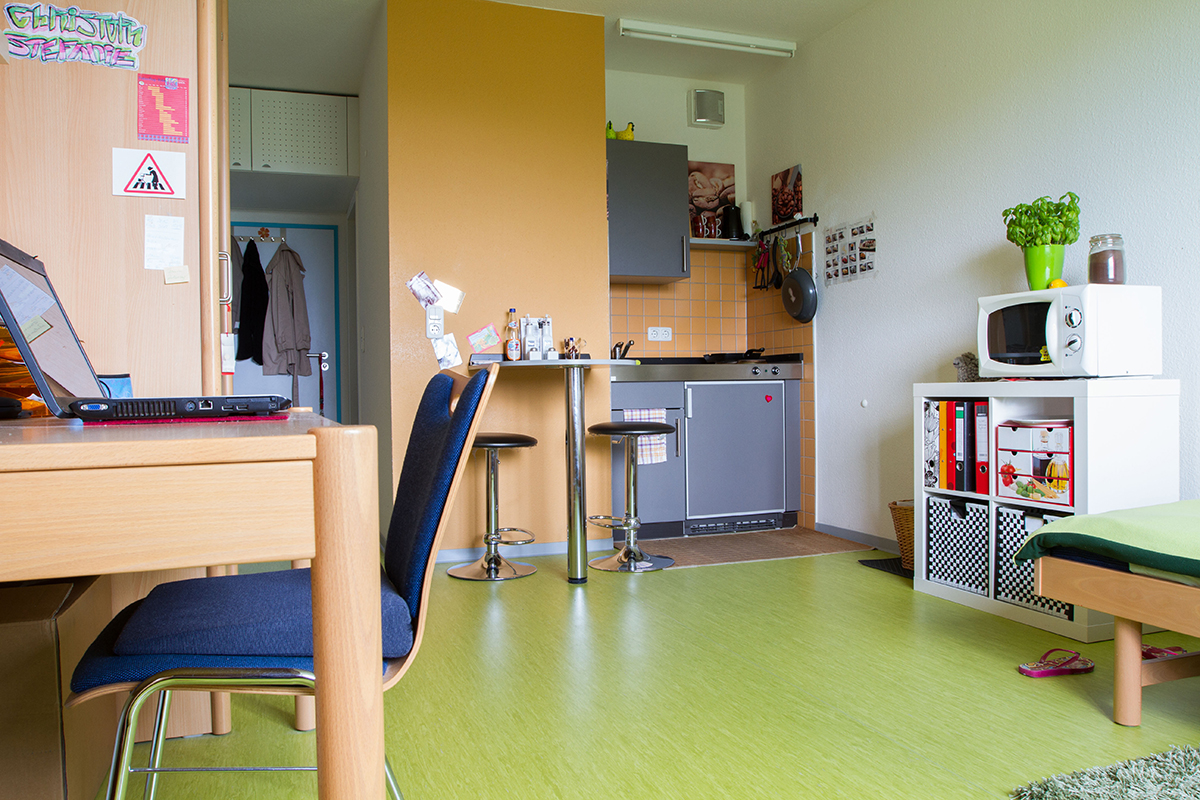 Ein kleiner, hellgrüner Raum mit Küchenzeile, Arbeitsplatz und Schlafbereich. Die Küche hat einen Herd, eine Mikrowelle und einen Kühlschrank. Der Arbeitsplatz besteht aus einem Tisch mit Laptop und Stuhl. Ein Bett mit Büchern und einer Pflanze befindet sich in der Ecke.