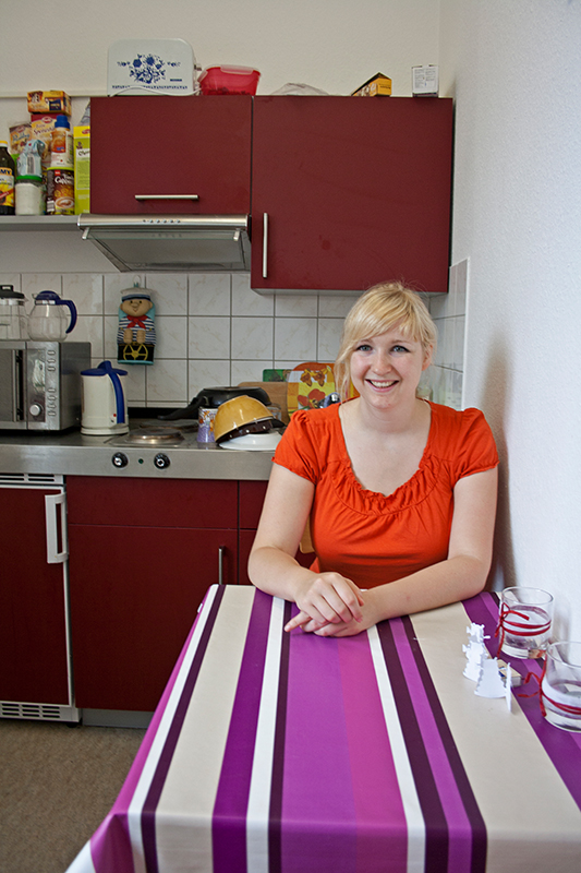 Eine Frau sitzt lächelnd an einem Tisch in einer Küche. Sie trägt ein oranges Oberteil. Der Tisch ist mit einer lila-weiß gestreiften Tischdecke bedeckt. Im Hintergrund sind Küchenschränke und Geräte zu sehen.