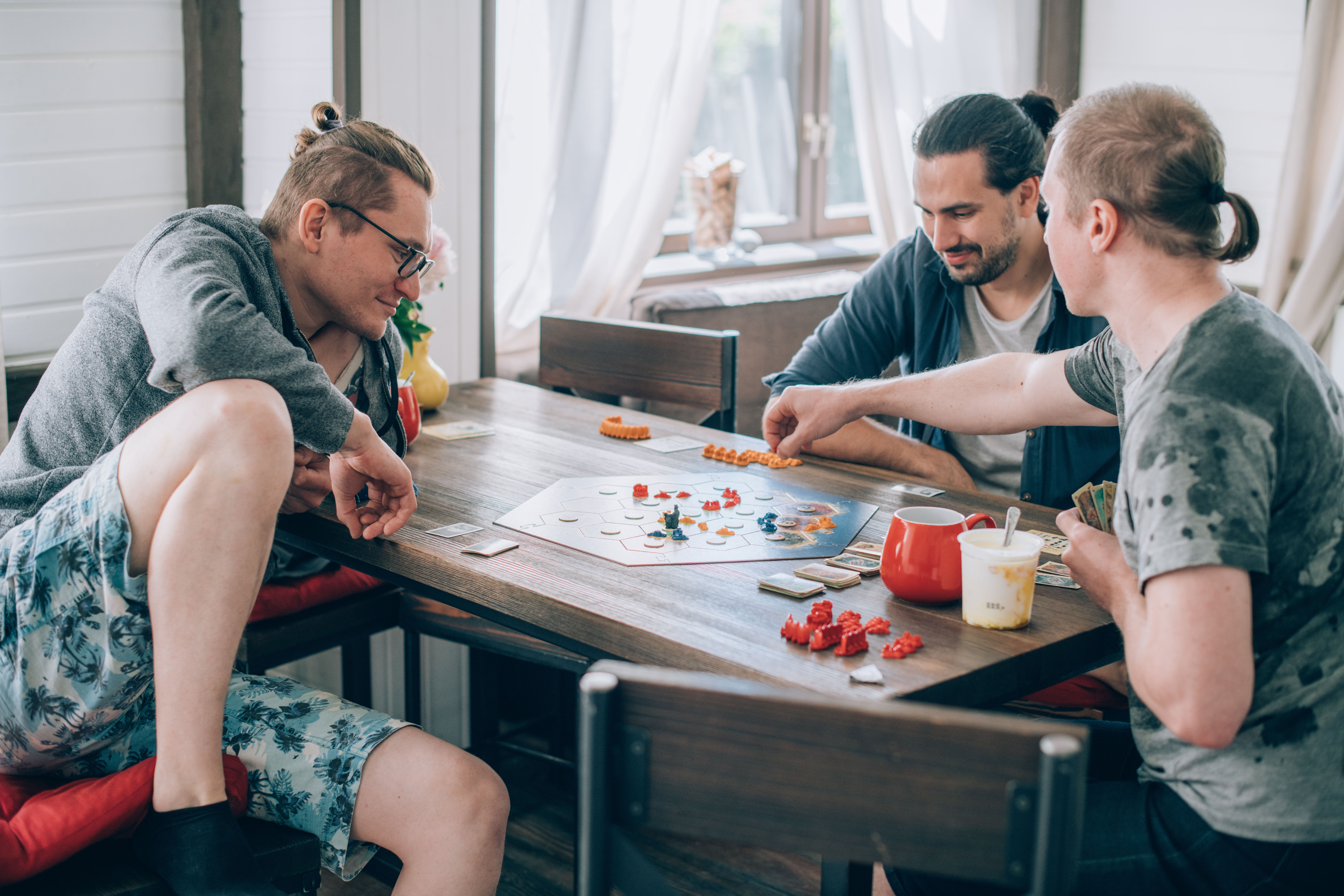 Drei Personen sitzen an einem Tisch und spielen ein Brettspiel. Sie wirken konzentriert und unterhalten sich dabei. Auf dem Tisch stehen Snacks und Getränke. Der Raum ist hell und freundlich mit großen Fenstern im Hintergrund.