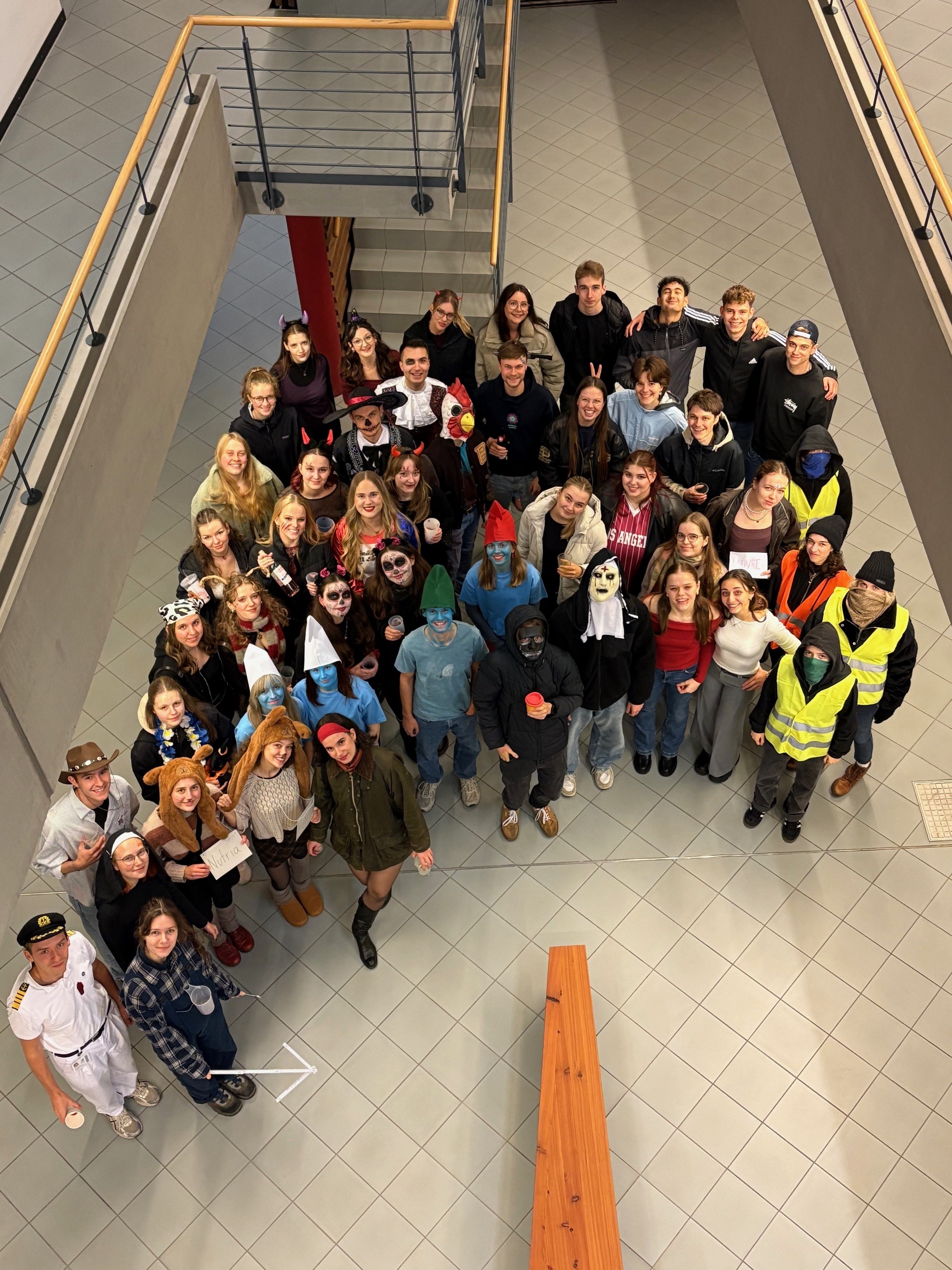 Eine große Gruppe von Menschen steht auf einer Treppe und lächelt in die Kamera. Die Personen sind unterschiedlich gekleidet, einige tragen Uniformen oder spezielle Kleidung. Die Szene wirkt fröhlich und gesellig, möglicherweise handelt es sich um eine Schulklasse oder eine Gruppe von Kollegen.