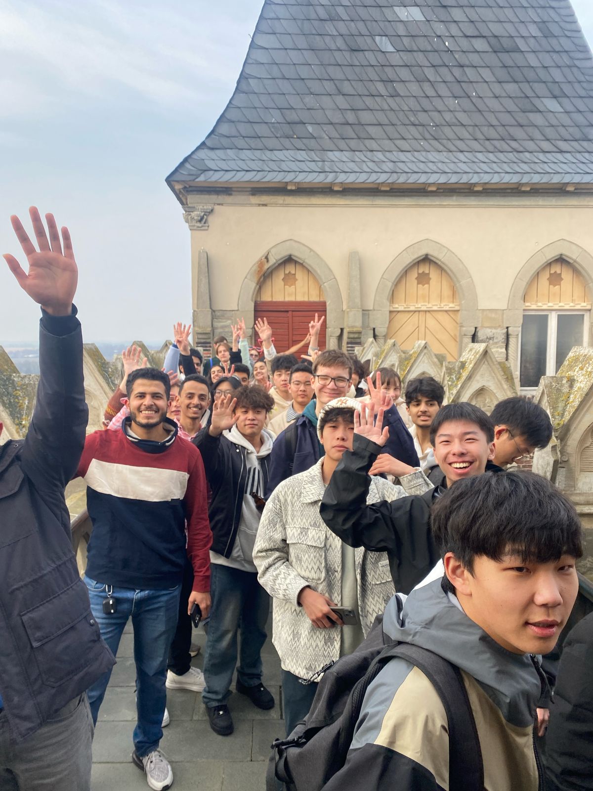 Eine Gruppe von Menschen steht vor einem Gebäude mit spitzem Dach und winkenden Händen. Die Personen lächeln und wirken fröhlich. Es scheint sich um eine Gruppe von Studierenden oder Teilnehmern einer Veranstaltung zu handeln, die sich für ein Gruppenfoto versammelt haben.