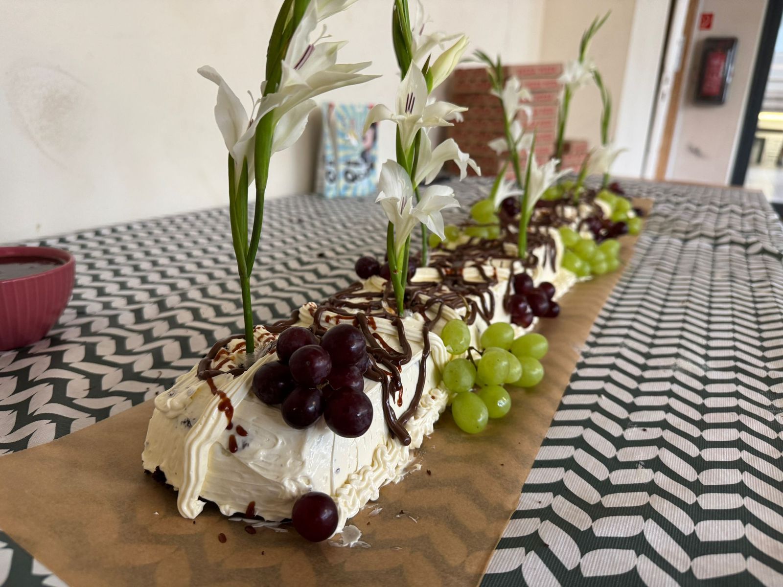 Eine Reihe von kleinen Kuchenstücken, die auf braunem Papier liegen. Jedes Stück ist mit weißer Creme, Trauben und Schokoladenstreuseln verziert. Dazu gibt es weiße Blumen als Deko. Die Kuchenstücke sind auf einem Tisch mit gemustertem Tischtuch angeordnet.